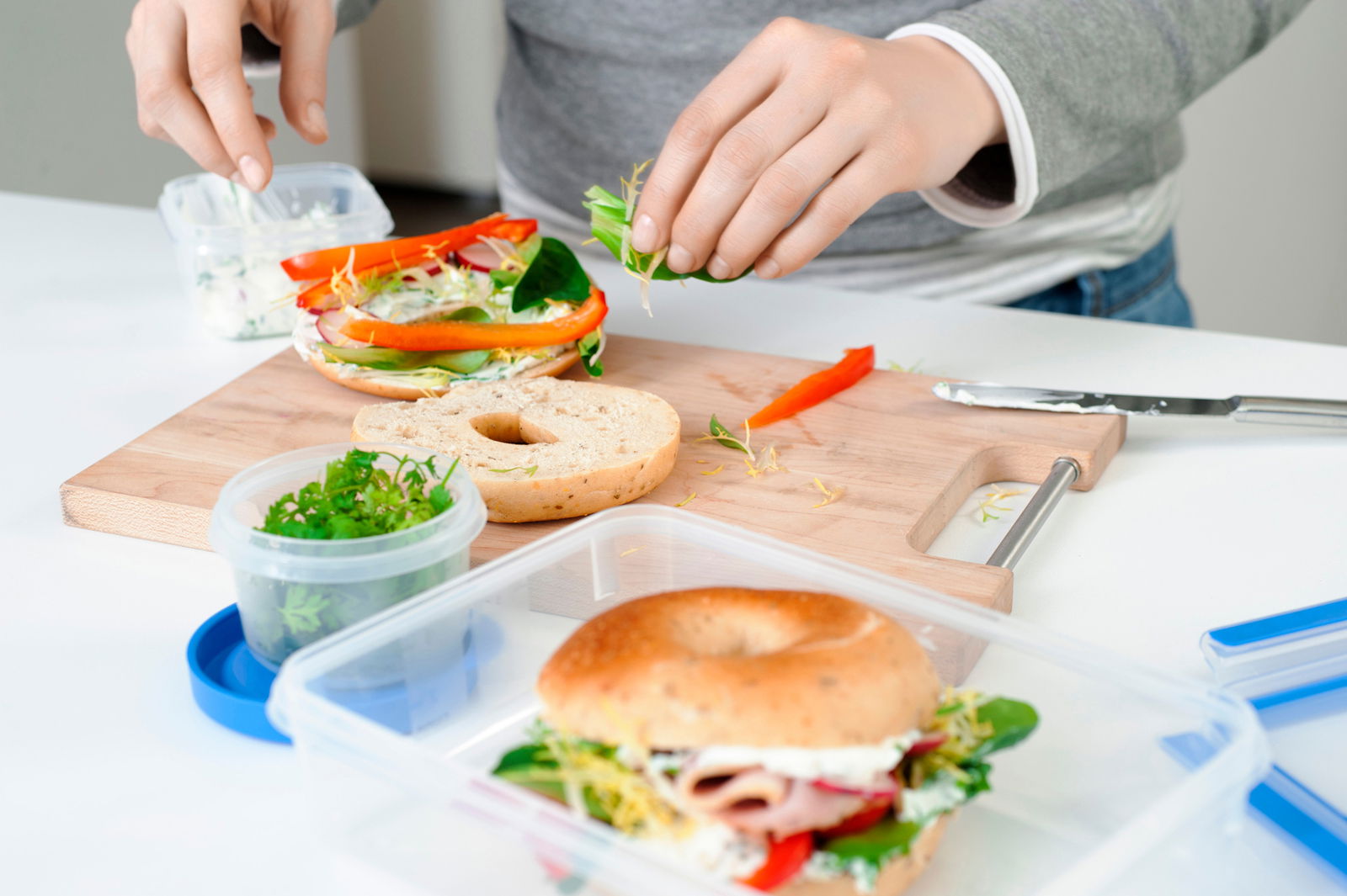 Frischhaltedose mit frischen Kräutern auf einem Tisch, im Hintergrund ein belegter Bagel auf einem Schneidebrett, Perspektive von oben