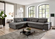 Großes graues Stoff-Wohnlandschaft-Ecksofa mit Ottomane links und breiten Armlehnen im hellen Wohnzimmer, Frontansicht in leichter Schrägperspektive von vorn