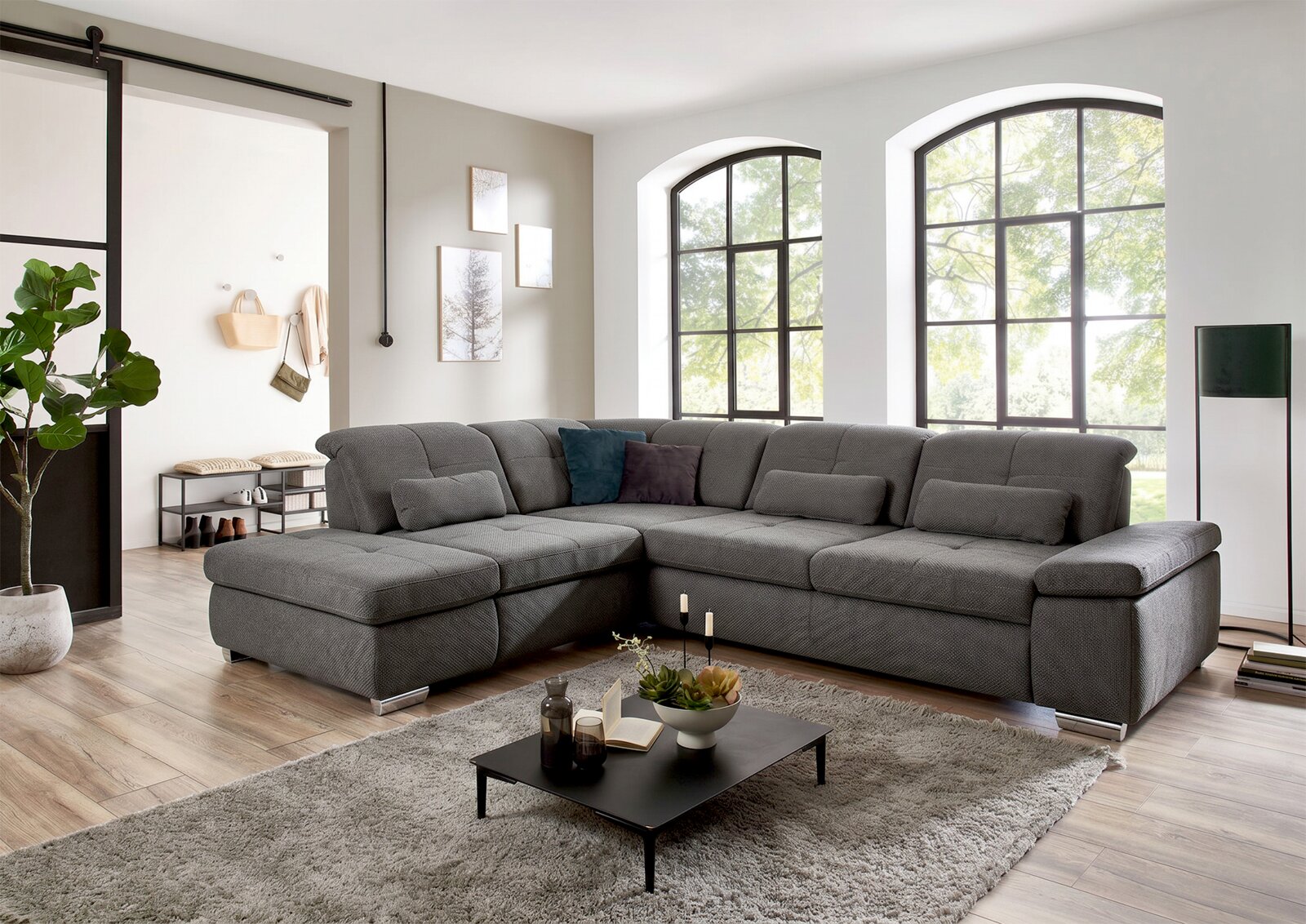 Großes graues Stoff-Wohnlandschaft-Ecksofa mit Ottomane links und breiten Armlehnen im hellen Wohnzimmer, Frontansicht in leichter Schrägperspektive von vorn