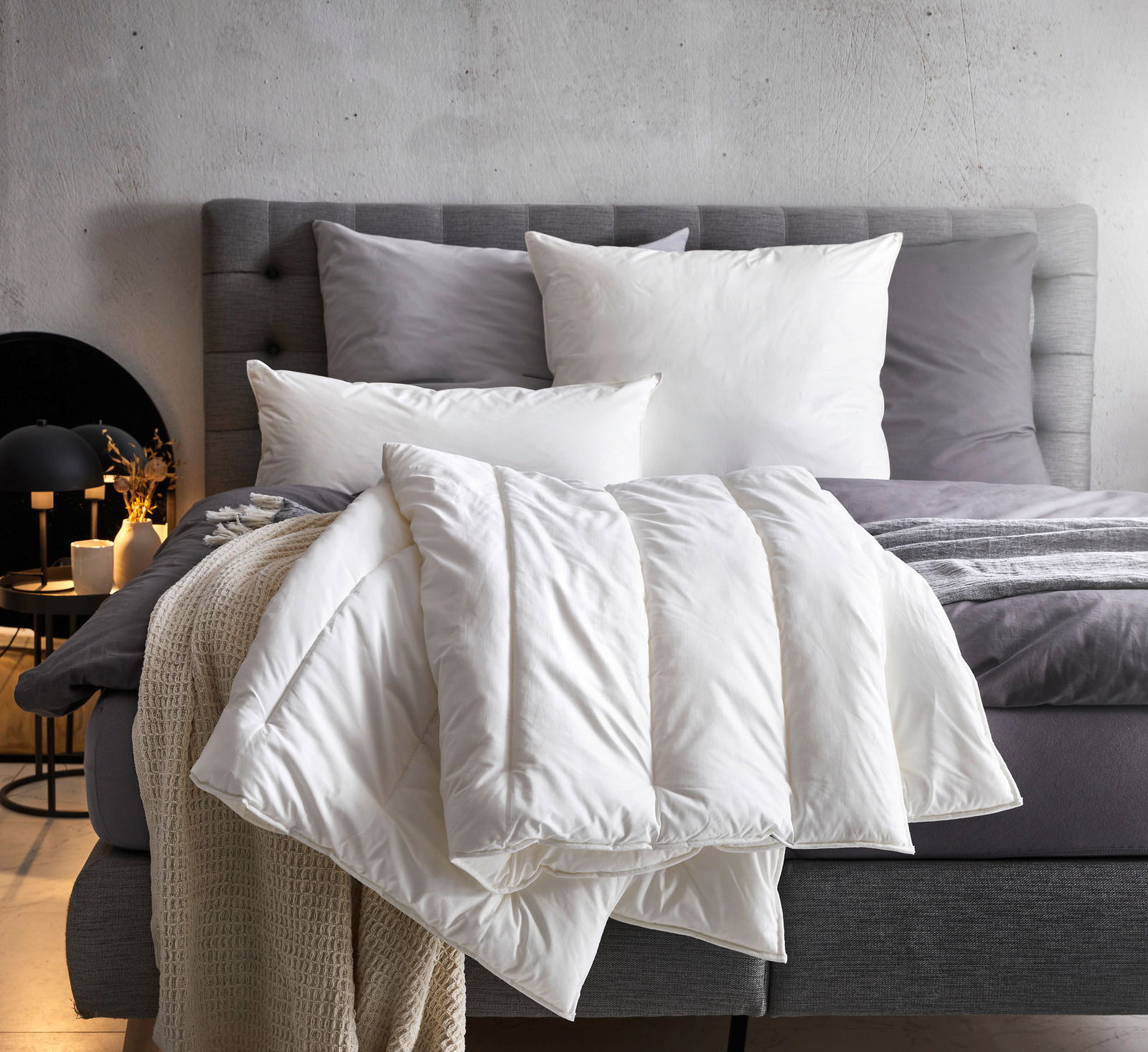 Gemütliches Solobett Vegan Dream 135x200 mit grauem Kopfteil, weißen und grauen Kissen sowie einer weißen Decke, seitliche Perspektive