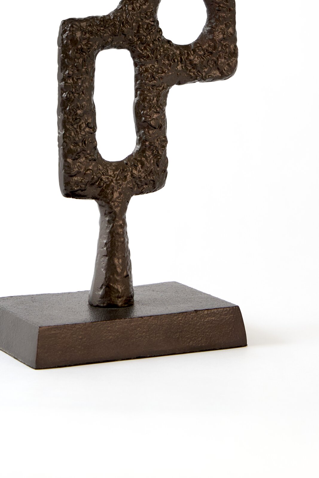 Abstract donkerbruin metalen sculptuur met ruwe textuur en geometrische openingen op een rechthoekige sokkel, schuin van voren gezien tegen een witte achtergrond