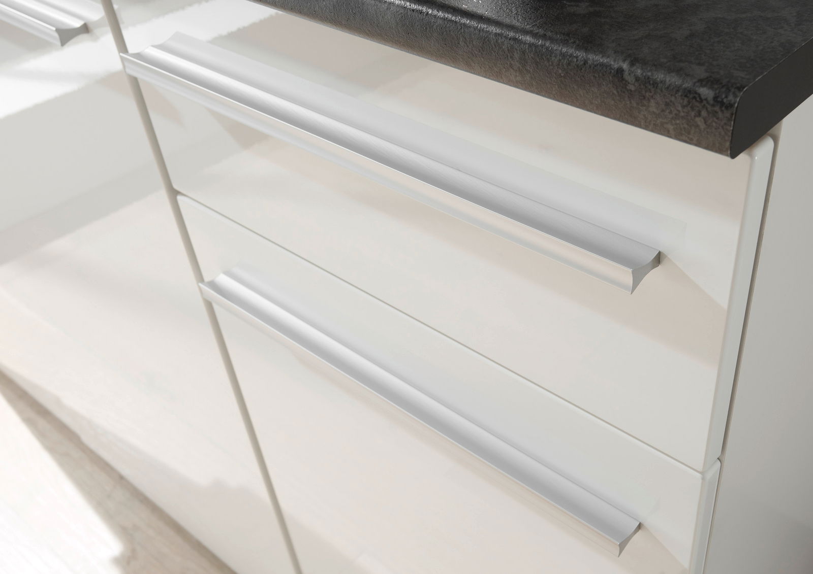 Nahaufnahme von weißen Küchenschubladen mit modernen, silbernen Griffen und einer dunklen Arbeitsplatte von oben betrachtet.