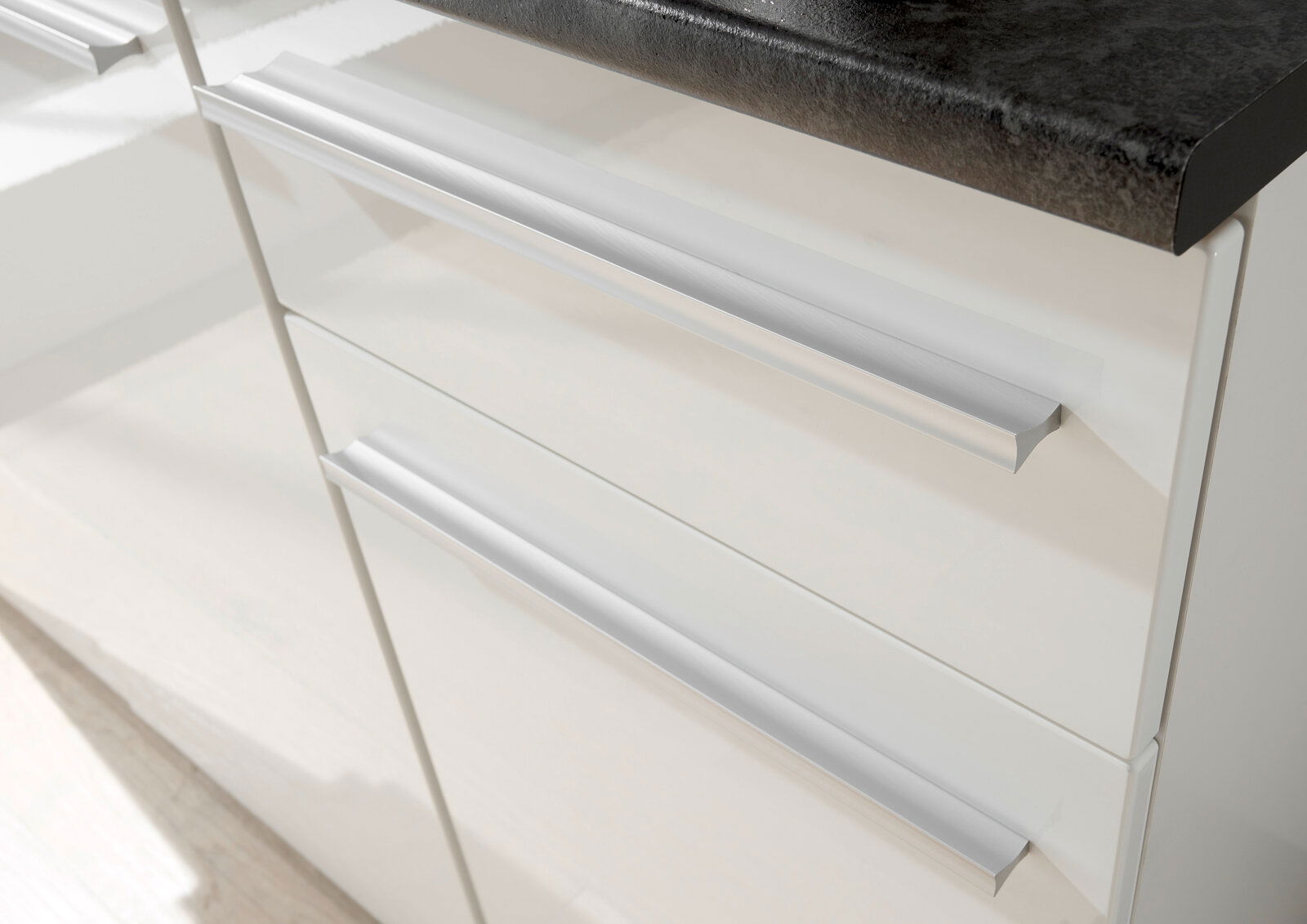 Nahaufnahme von weißen Küchenschubladen mit modernen, silbernen Griffen und einer dunklen Arbeitsplatte von oben betrachtet.