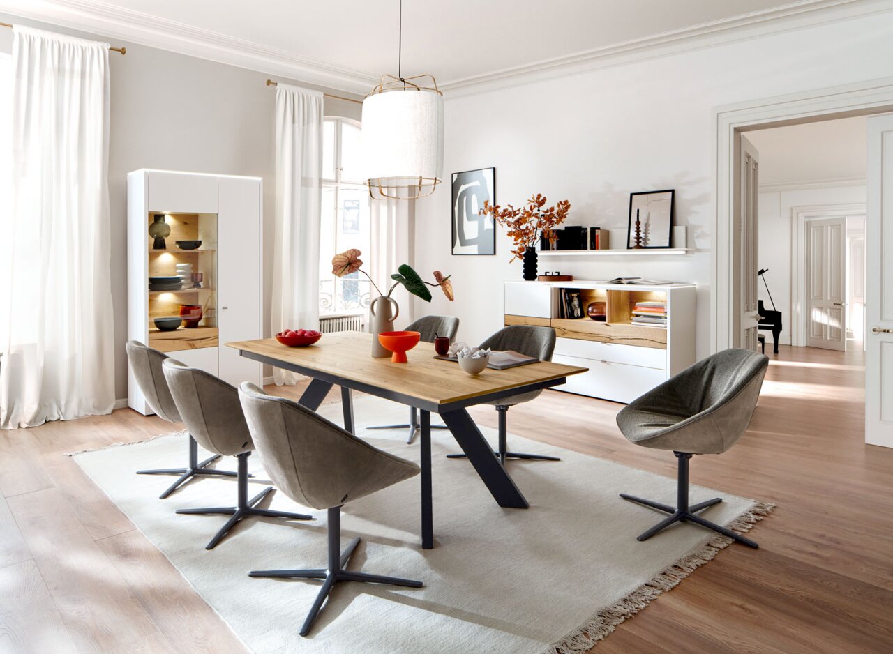 Venjakob Esstisch ET651 LEO Moderner Essbereich mit rechteckigem Esstisch aus Holz mit schwarzem Metallgestell und vier grauen Polsterstühlen, im Hintergrund weiße Vitrine und Sideboard; Perspektive: leicht schräg von vorne auf Tisch und Stühle.