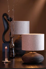Stimmungsvolle Nahaufnahme eines Lampenfußes mit rundem, gerilltem Design und einem hellen Lampenschirm, im Hintergrund eine dekorative Vase und eine brennende Kerze.