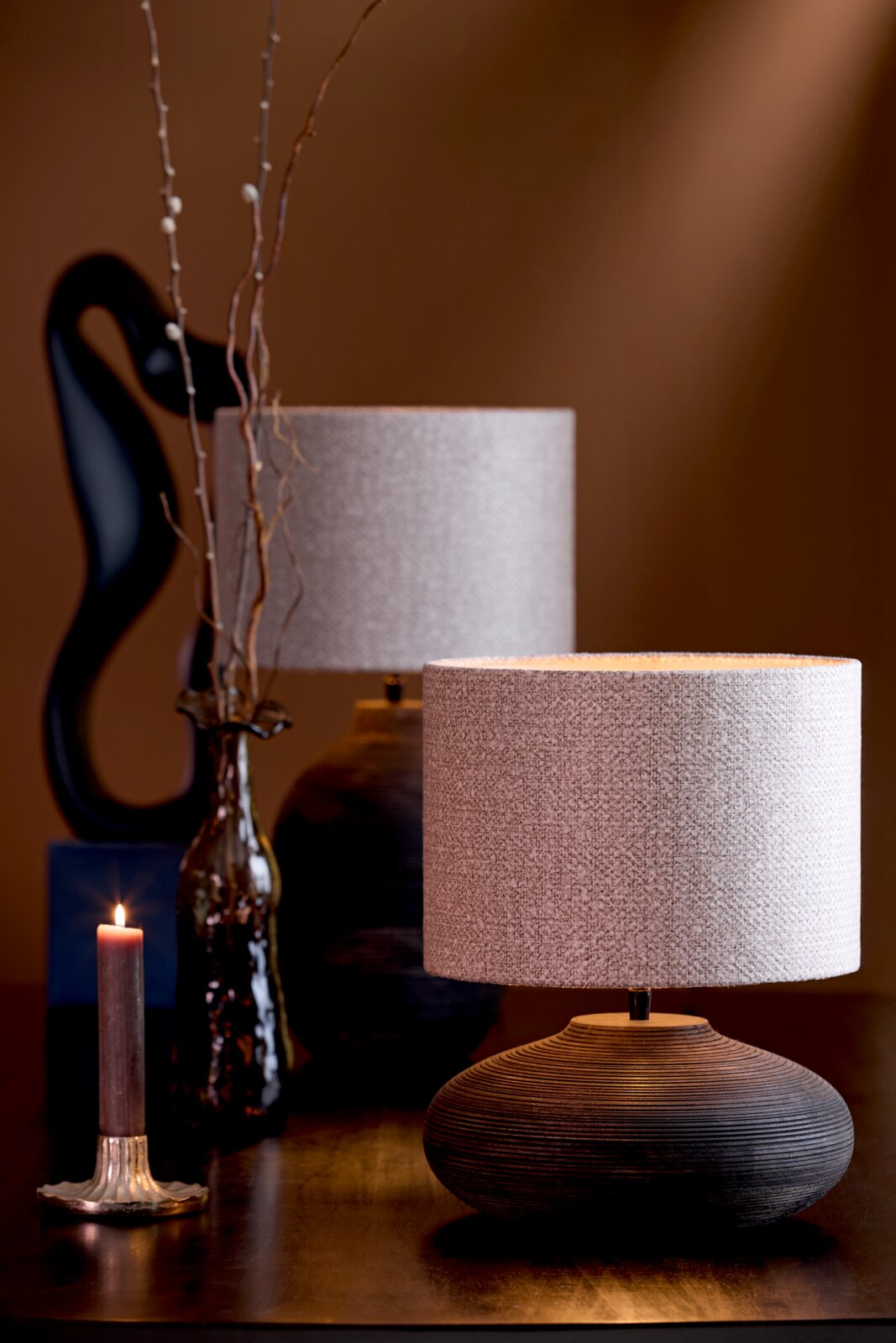 Stimmungsvolle Nahaufnahme eines Lampenfußes mit rundem, gerilltem Design und einem hellen Lampenschirm, im Hintergrund eine dekorative Vase und eine brennende Kerze.