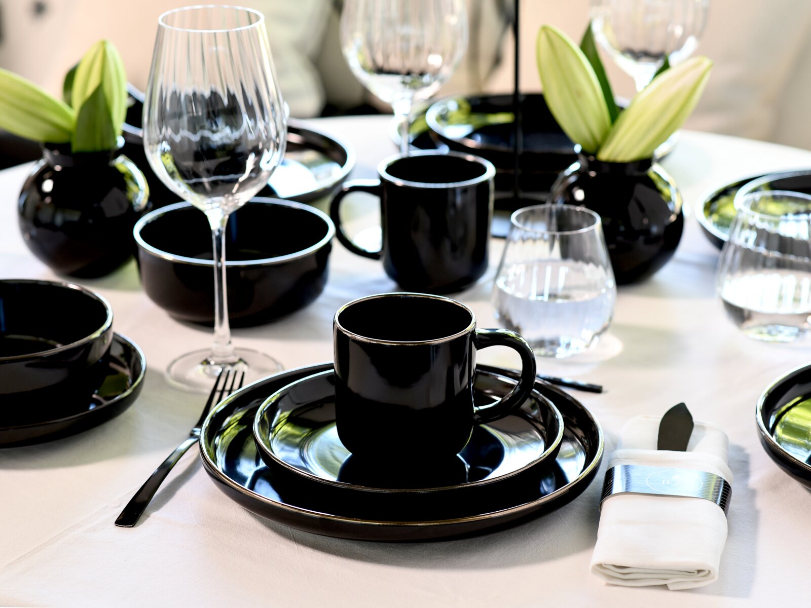 Elegant gedeckter Tisch mit schwarzem Geschirr (Tassen, Schalen und Teller), klaren Wein- und Wassergläsern sowie schwarzen Vasen mit grünen Knospen, aus leicht seitlicher Perspektive auf Augenhöhe aufgenommen.