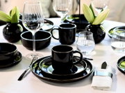 Elegant gedeckter Tisch mit schwarzem Geschirr (Tassen, Schalen und Teller), klaren Wein- und Wassergläsern sowie schwarzen Vasen mit grünen Knospen, aus leicht seitlicher Perspektive auf Augenhöhe aufgenommen.
