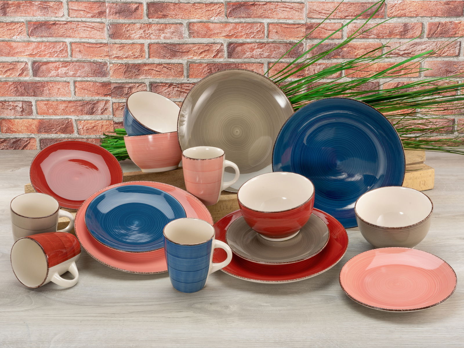 Kombiservice 16-tlg. in verschiedenen Farben, bestehend aus Tellern, Schalen und Tassen, auf einem Tisch vor einer Ziegelwand ausgestellt. Perspektive: Frontalansicht.
