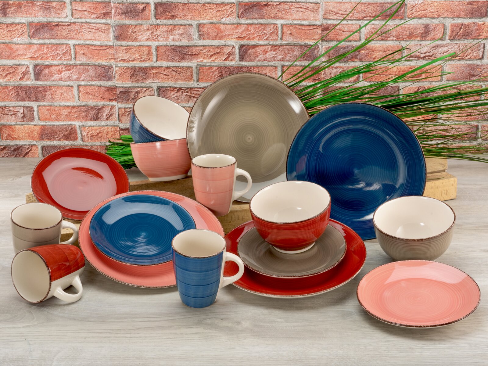 Serviesset van 16-delig in verschillende kleuren, bestaande uit borden, schalen en kopjes, uitgestald op een tafel voor een bakstenen muur. Perspectief: vooraanzicht.