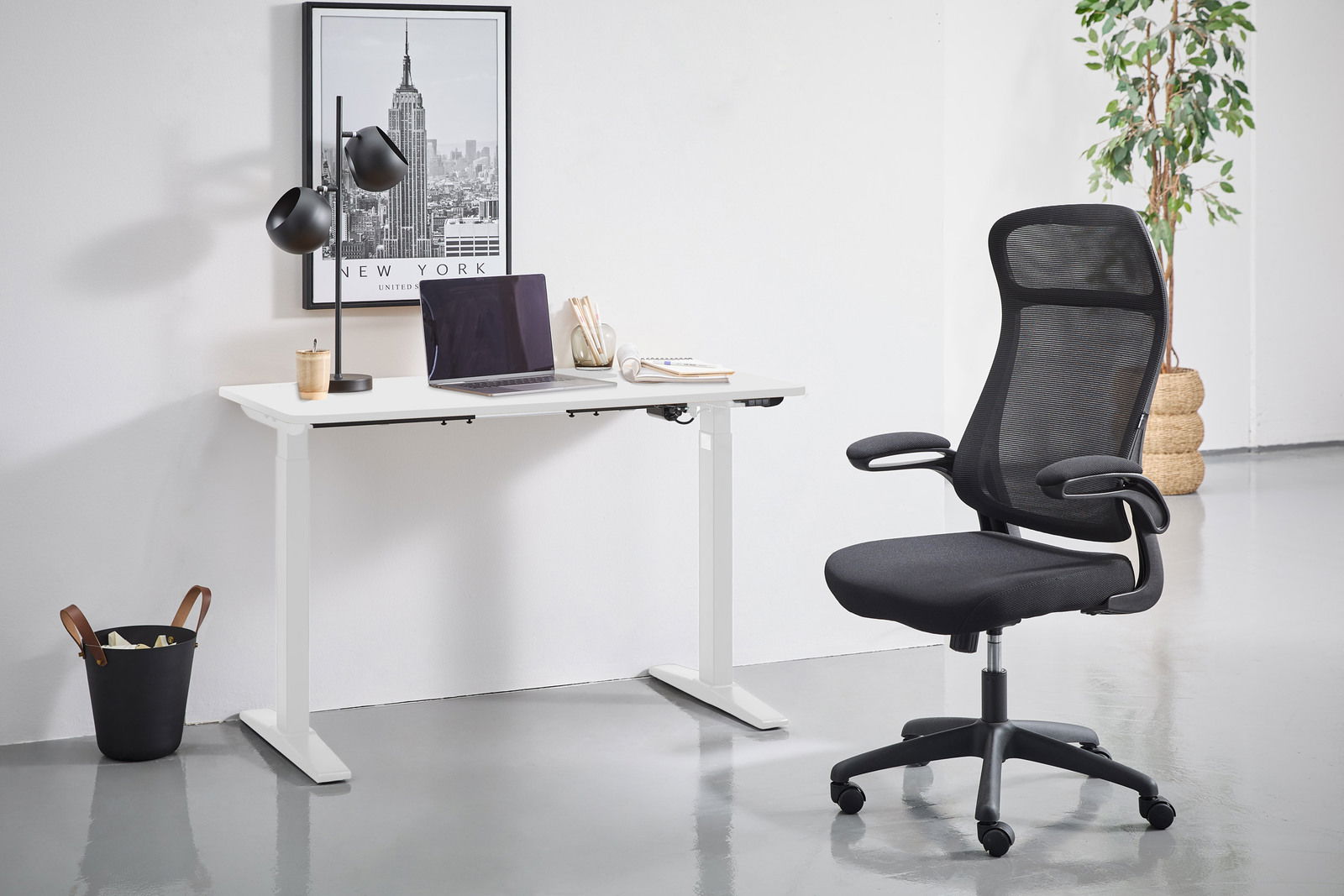 Moderner E-Schreibtisch in Weiß mit schwarzem Bürostuhl, seitliche Perspektive. Der Schreibtisch ist mit einer Lampe, einem Laptop und Schreibutensilien dekoriert. Im Hintergrund ein New York Poster und eine Pflanze.
