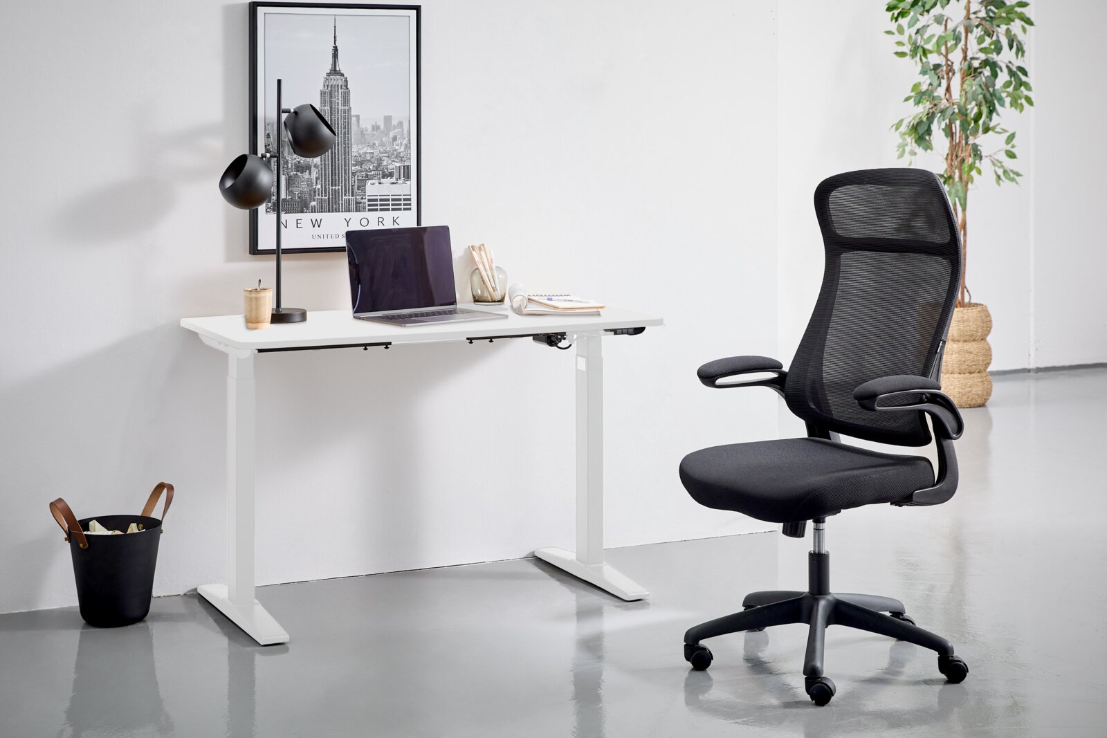 Moderner E-Schreibtisch in Weiß mit schwarzem Bürostuhl, seitliche Perspektive. Der Schreibtisch ist mit einer Lampe, einem Laptop und Schreibutensilien dekoriert. Im Hintergrund ein New York Poster und eine Pflanze.
