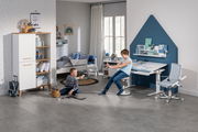 Moderner Schreibtisch in einem Kinderzimmer, seitliche Perspektive, mit ergonomischem Stuhl und Regal, umgeben von spielenden Kindern.