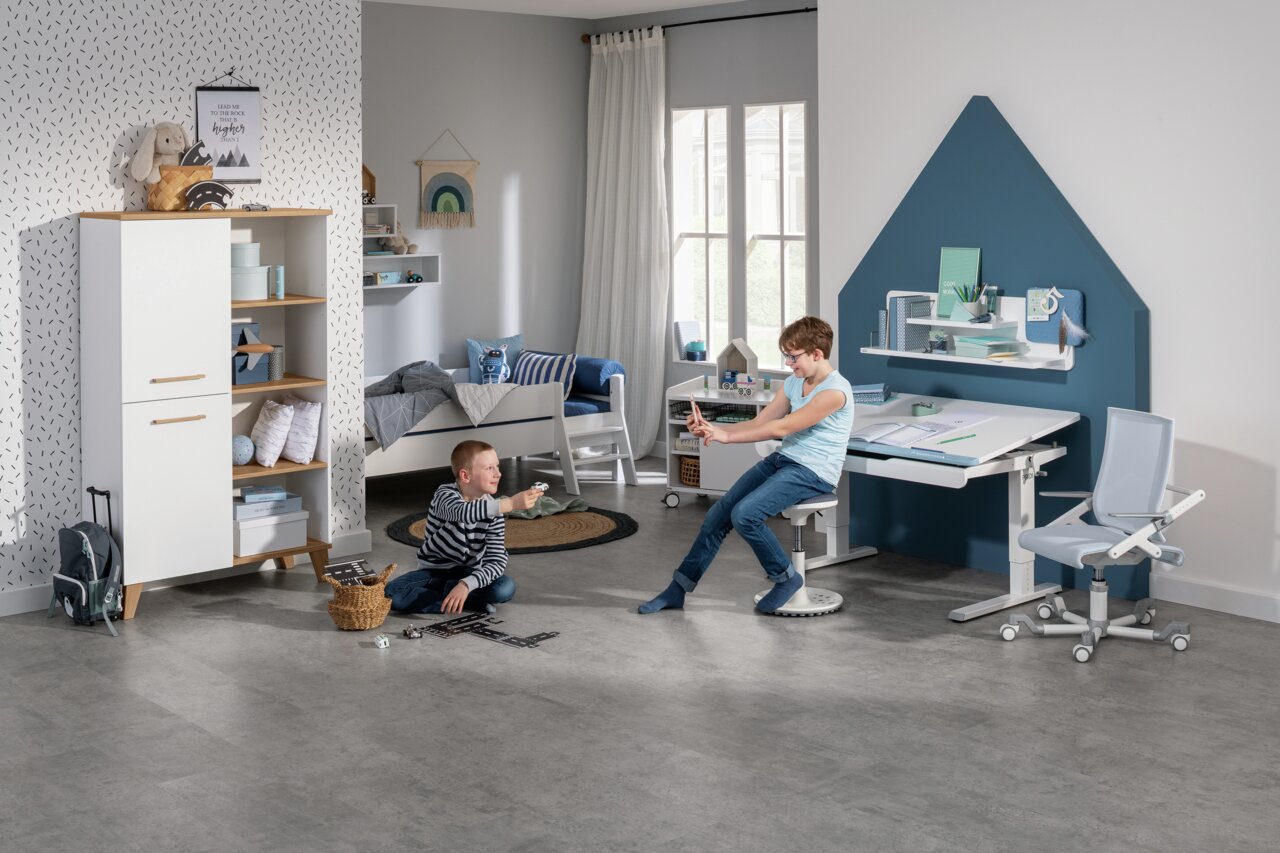 Modern bureau in een kinderkamer, gezien vanaf de zijkant, met een ergonomische stoel en een boekenkast, omringd door spelende kinderen.