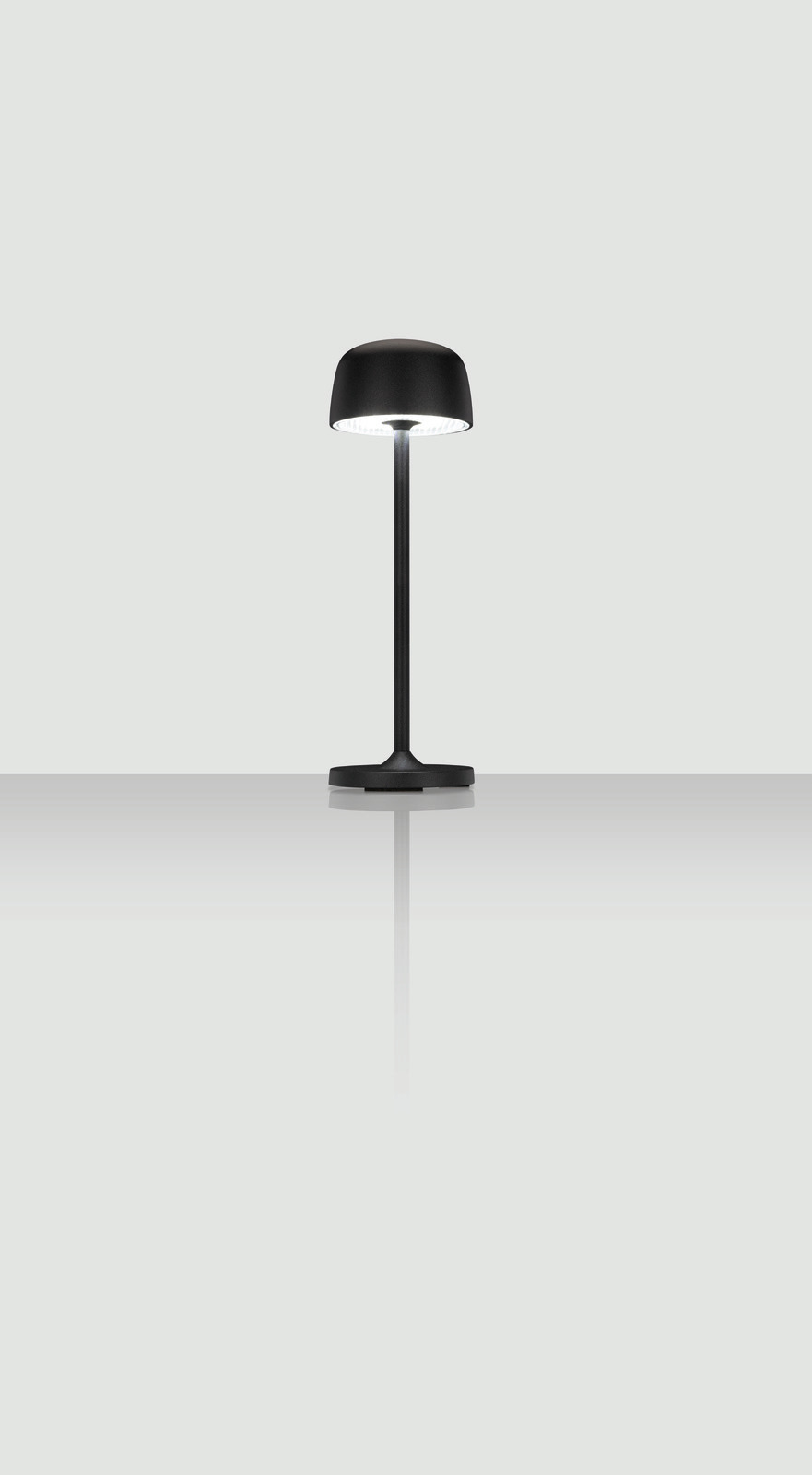 Moderne schwarze Akku-Tischleuchte mit rundem Schirm, frontal betrachtet auf reflektierender Oberfläche.
