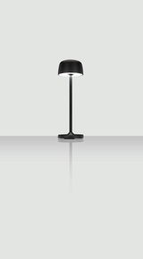 Moderne schwarze Akku-Tischleuchte mit rundem Schirm, frontal betrachtet auf reflektierender Oberfläche.
