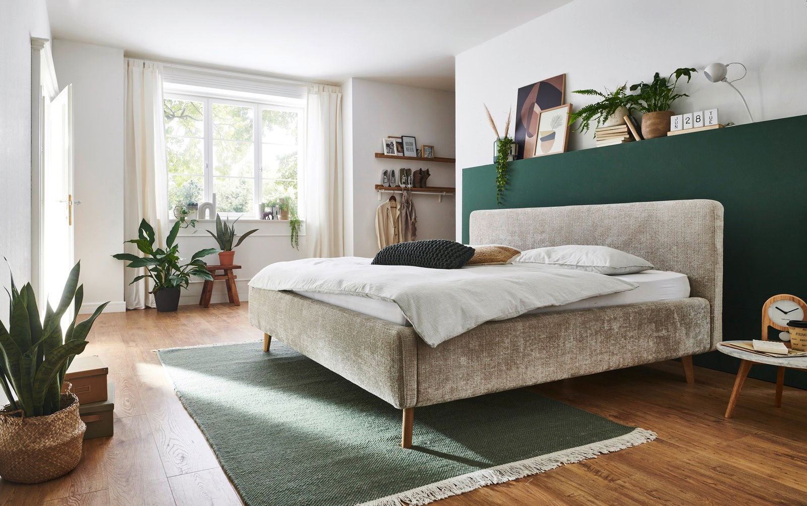 Modernes Polsterbett in einem hellen Schlafzimmer, seitliche Perspektive, mit grüner Wand und Pflanzen dekoriert.