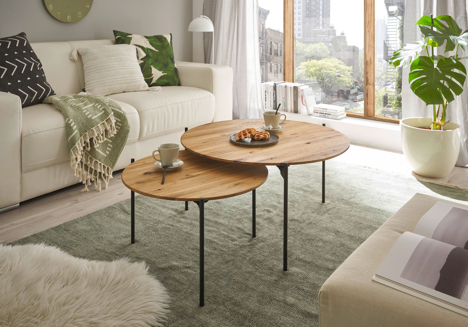 Zwei runde Beistelltische aus Holz mit schwarzen Metallbeinen, im Wohnzimmer vor einem Sofa, mit Blick auf ein großes Fenster.