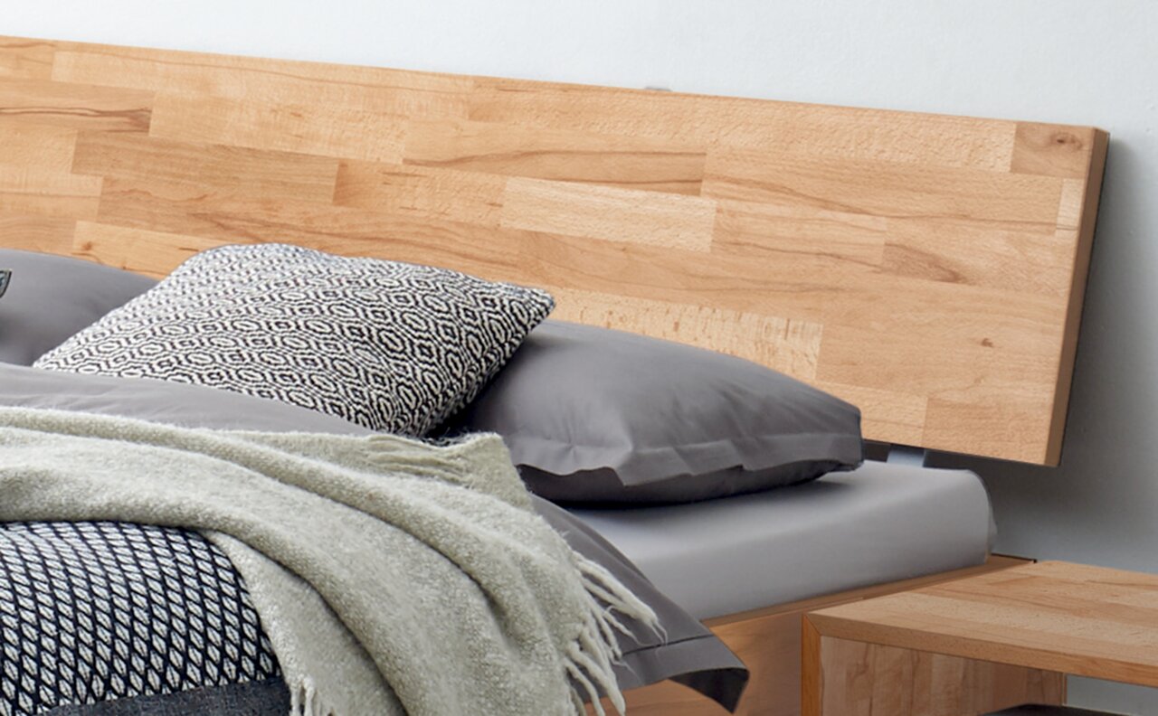 Holzbettgestell mit Kopfteil aus hellem Holz, graue Bettwäsche und gemustertes Kissen, seitliche Perspektive