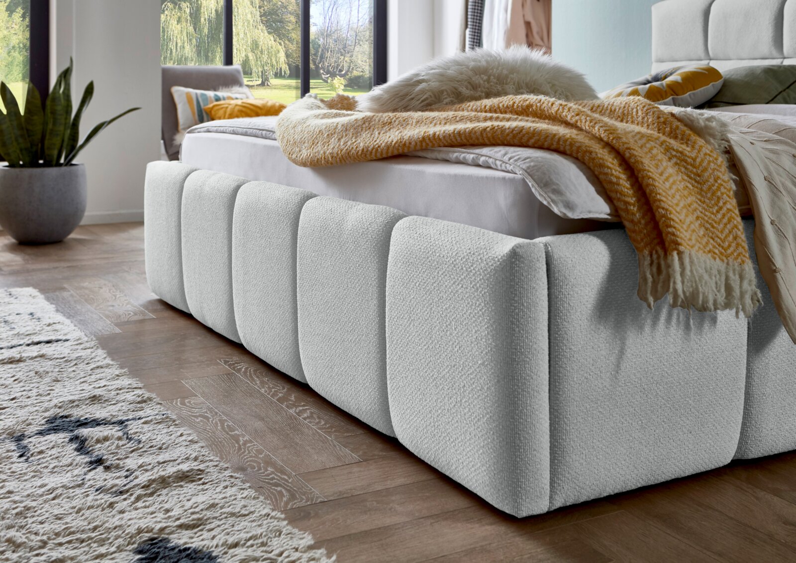 Close-up van een modern gestoffeerd bed in een lichte kamer met grote ramen, gefotografeerd vanaf de zijkant. Het bed is bekleed met een zachte, gestructureerde stof en versierd met een gele deken.
