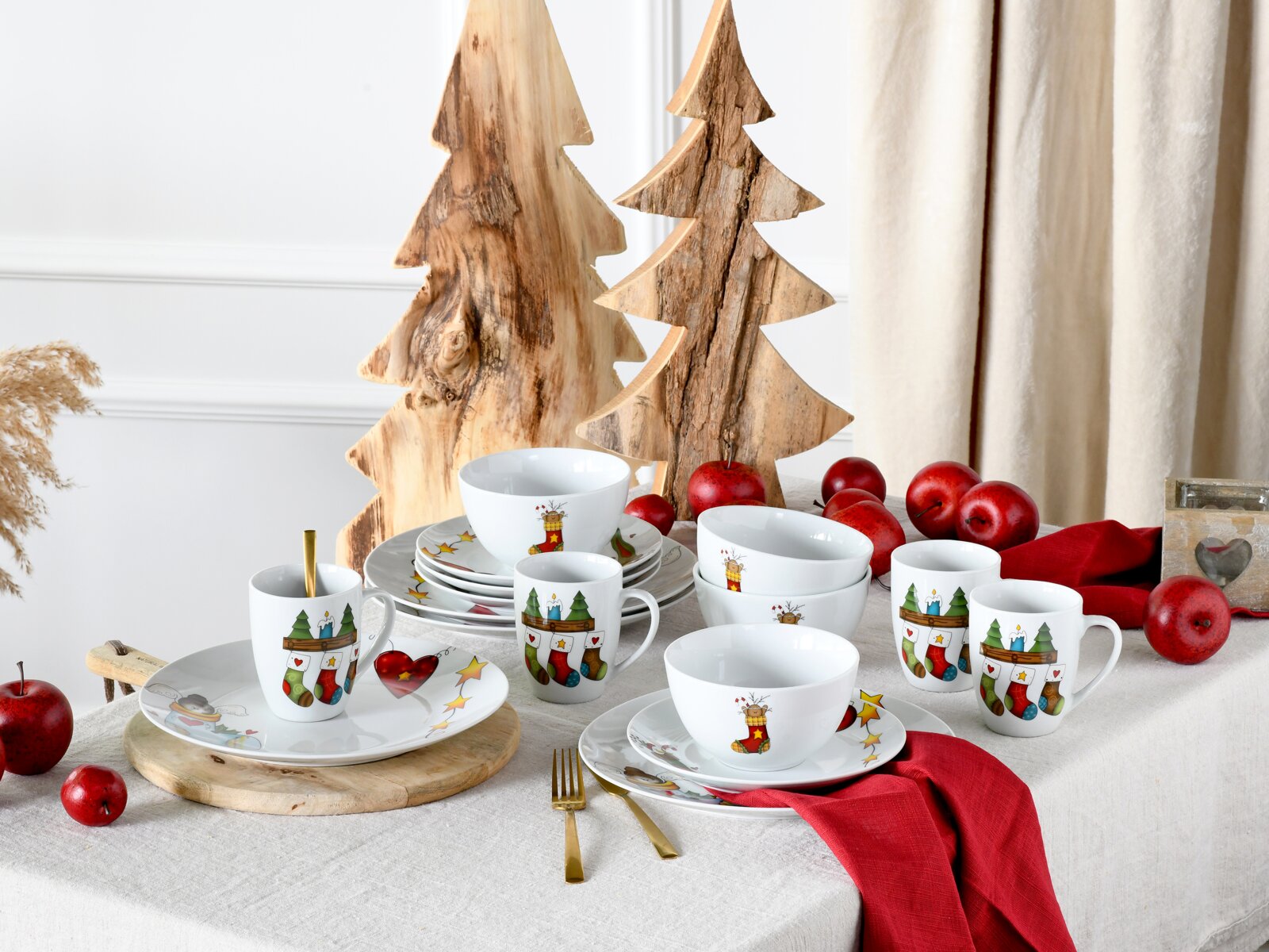 Weihnachtliches Porzellan-Geschirrset mit Tassen, Schalen und Tellern mit bunten Strumpf- und Sternmotiven auf gedecktem Tisch, dekoriert mit roten Äpfeln und Holz-Tannenbäumen, Aufnahme in leicht schräger Frontansicht auf Augenhöhe