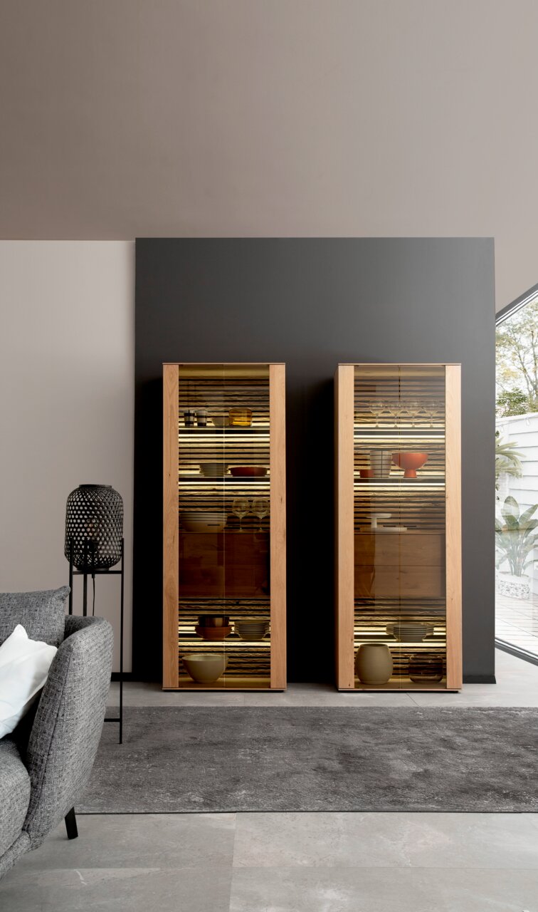 Zwei moderne Vitrinen aus Holz mit Glastüren, frontal fotografiert, gefüllt mit dekorativen Objekten, in einem stilvollen Wohnzimmer.