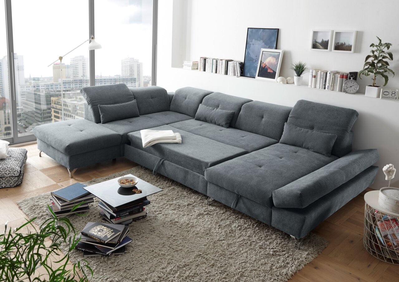 Großes, graues Ecksofa in einem modernen Wohnzimmer mit Blick auf die Stadt durch große Fenster. Perspektive von schräg oben.