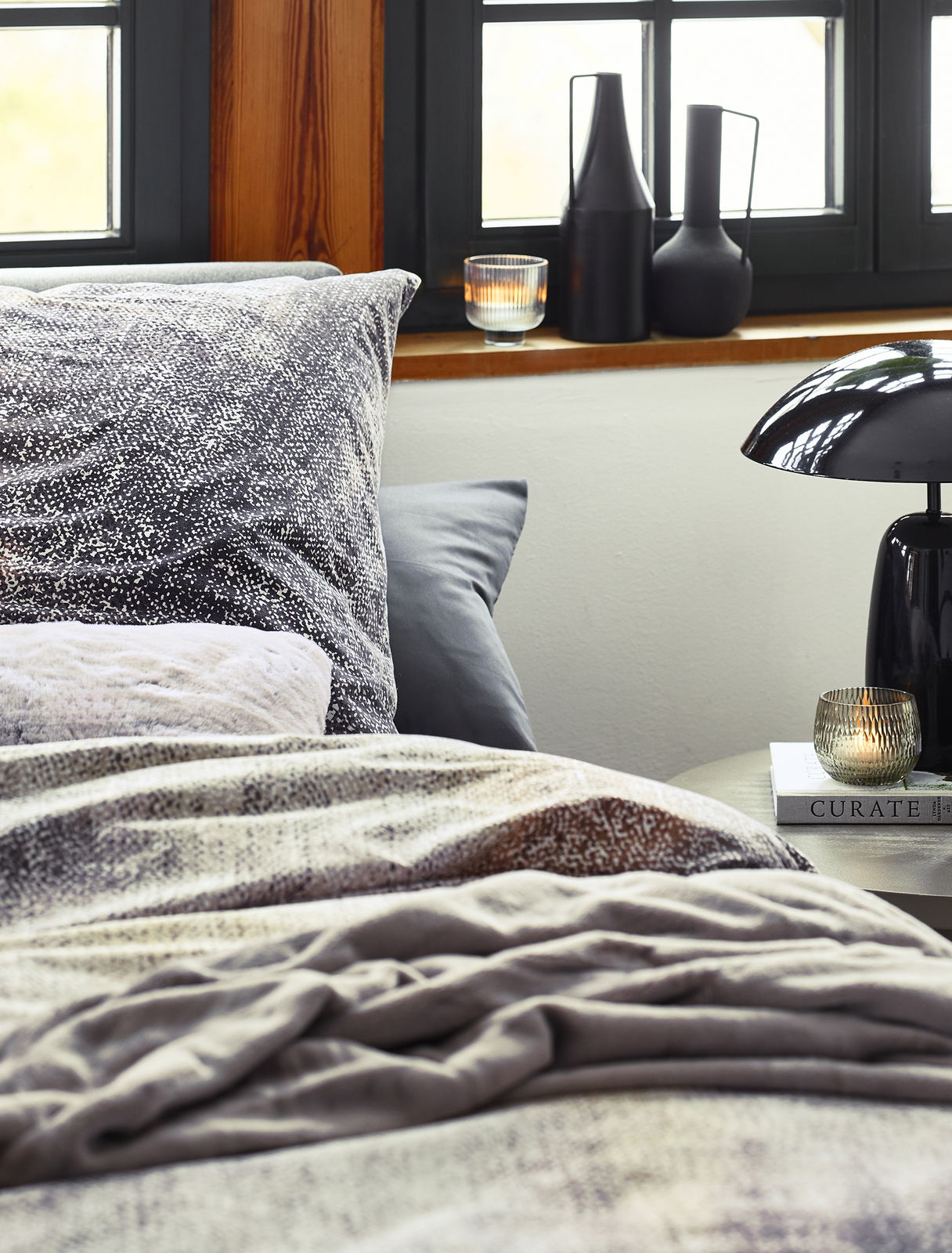 Gemütliche Bettwäsche in einem modernen Schlafzimmer mit Fenster im Hintergrund, dekorativen Vasen und einer Lampe auf dem Nachttisch, Perspektive von der Bettkante.