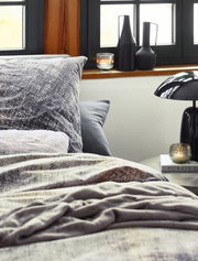 Gemütliche Bettwäsche in einem modernen Schlafzimmer mit Fenster im Hintergrund, dekorativen Vasen und einer Lampe auf dem Nachttisch, Perspektive von der Bettkante.