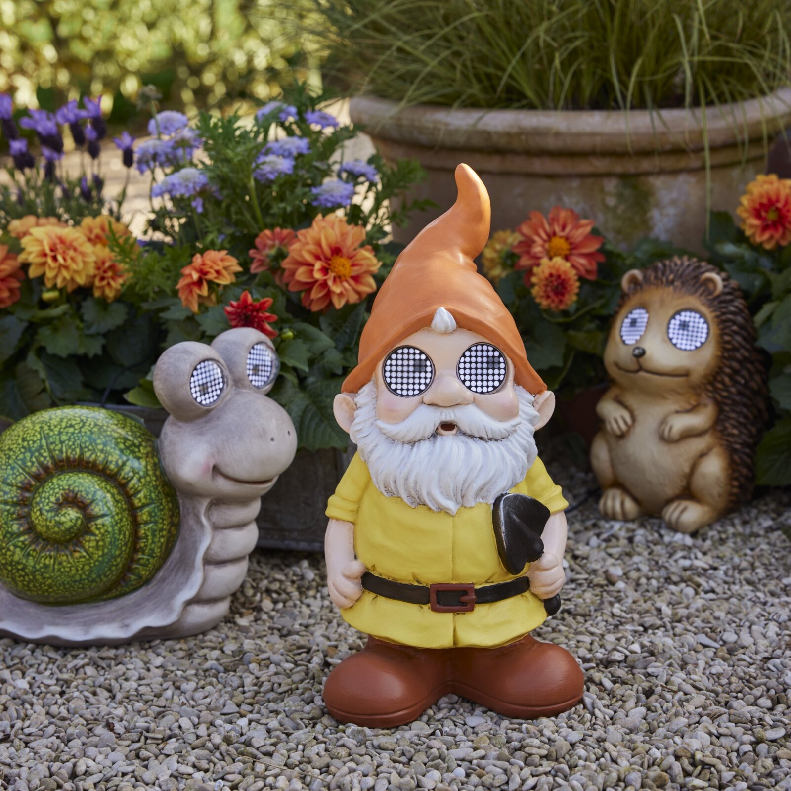 Gartenfigur eines Solarzwergs aus Polyresin mit orangefarbener Mütze und gelbem Anzug, umgeben von einer Schnecke und einem Igel mit leuchtenden Augen, auf einem Kiesweg vor blühenden Pflanzen. Frontalansicht.