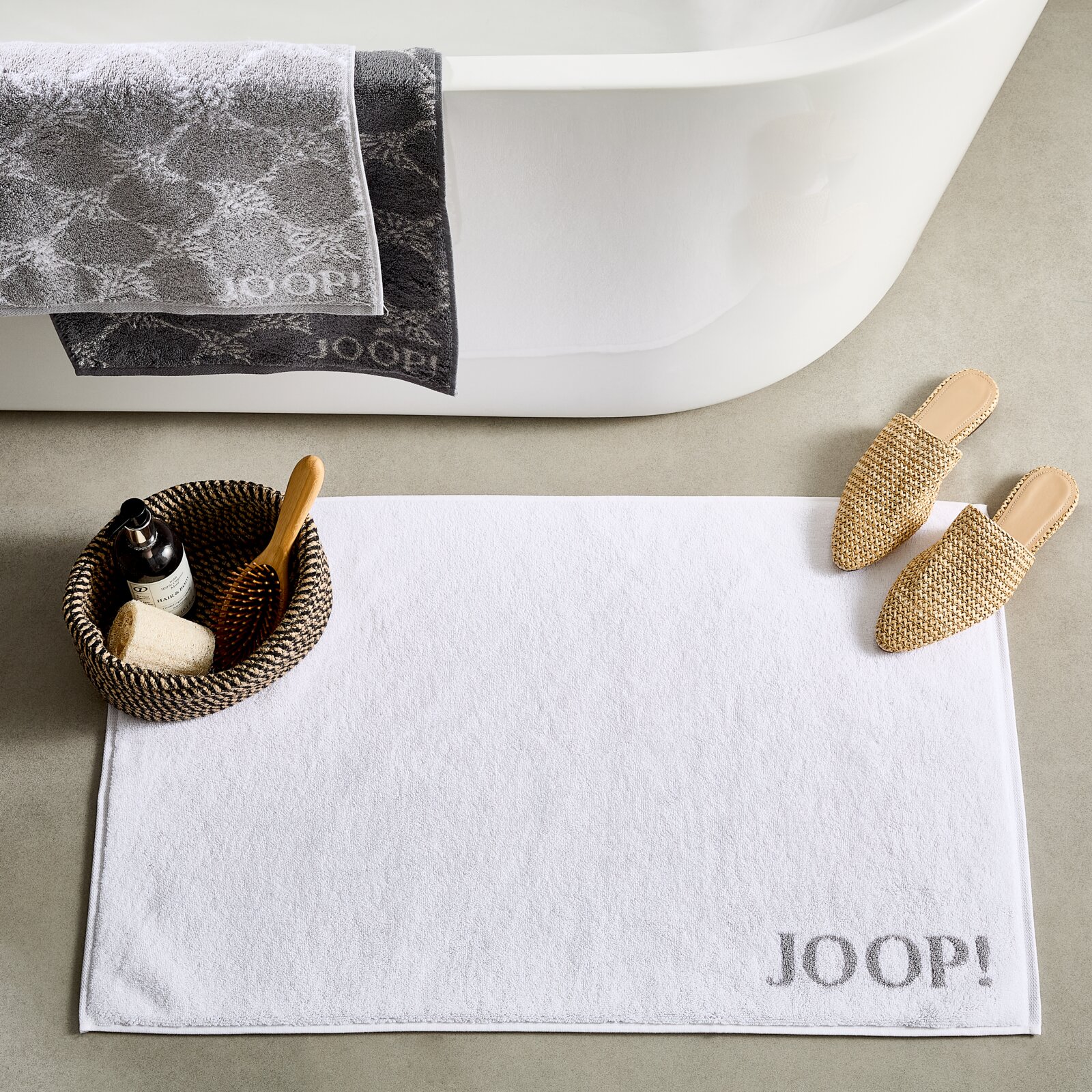 Weiße Badematte mit 'JOOP!' Schriftzug, platziert vor einer Badewanne, mit dekorativen Elementen wie einem Korb und Hausschuhen. Draufsicht.