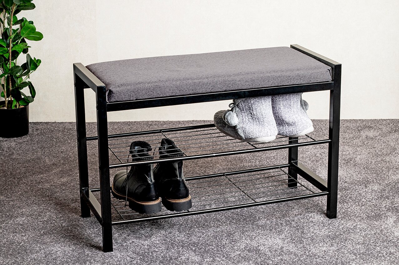 Schwarze Schuhbank aus Metall mit grauem Sitzpolster und zwei Ablagegittern, mit Schuhen darunter, aus leicht schräger Frontansicht auf grauem Teppich vor heller Wand.