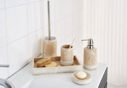 Beiges Badezimmer-Accessoire-Set in Steinoptik mit Seifenspender, Zahnputzbecher, WC-Bürstenhalter, Ablageschale und Seifenschale auf weißem Waschbecken, perspektivisch aus leicht schräger Frontansicht