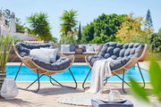 Zwei gemütliche Relaxsessel aus Rattan mit dunkelgrauen Kissen, aufgestellt auf einer Terrasse mit Blick auf einen Pool und umgeben von grünen Pflanzen.