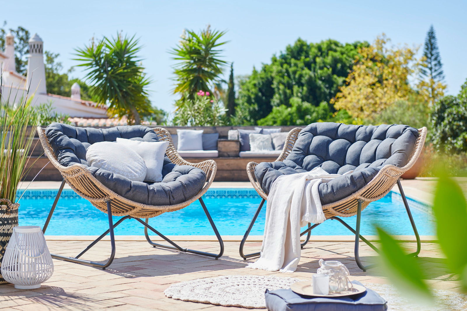 Zwei gemütliche Relaxsessel aus Rattan mit dunkelgrauen Kissen, aufgestellt auf einer Terrasse mit Blick auf einen Pool und umgeben von grünen Pflanzen.
