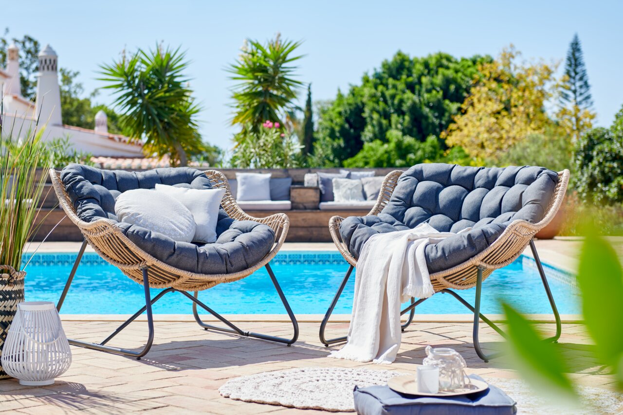 Ambia Garden Relaxfauteuil CHAMBERY Twee comfortabele rotanfauteuils met donkergrijze kussens, geplaatst op een terras met uitzicht op een zwembad en omringd door groene planten.