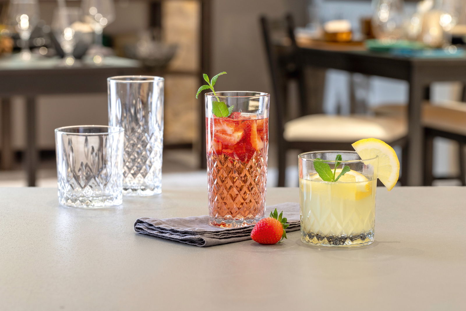 Vier elegante Gläser mit Diamantschliff-Design, gefüllt mit Getränken und dekoriert mit Minze und Zitrone, auf einem Tisch in einem stilvollen Esszimmer.