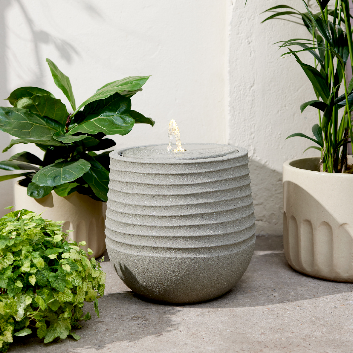 Moderner Brunnen aus recyceltem Kunststoff mit wellenförmigem Design, umgeben von Pflanzen, aus seitlicher Perspektive fotografiert.