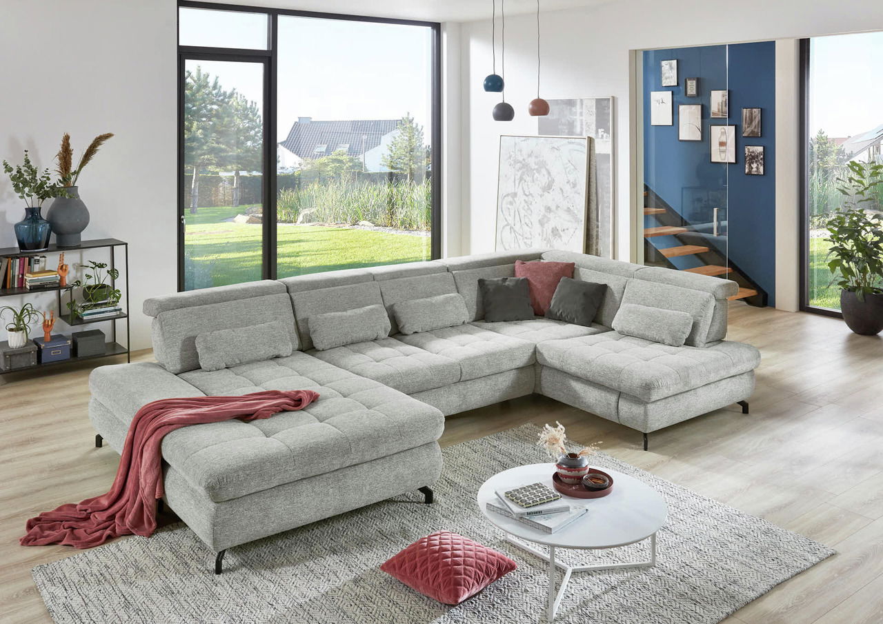 Graue Polsterrundecke in modernem Wohnzimmer mit großen Fenstern, dekoriert mit Kissen und Decke, seitliche Perspektive.