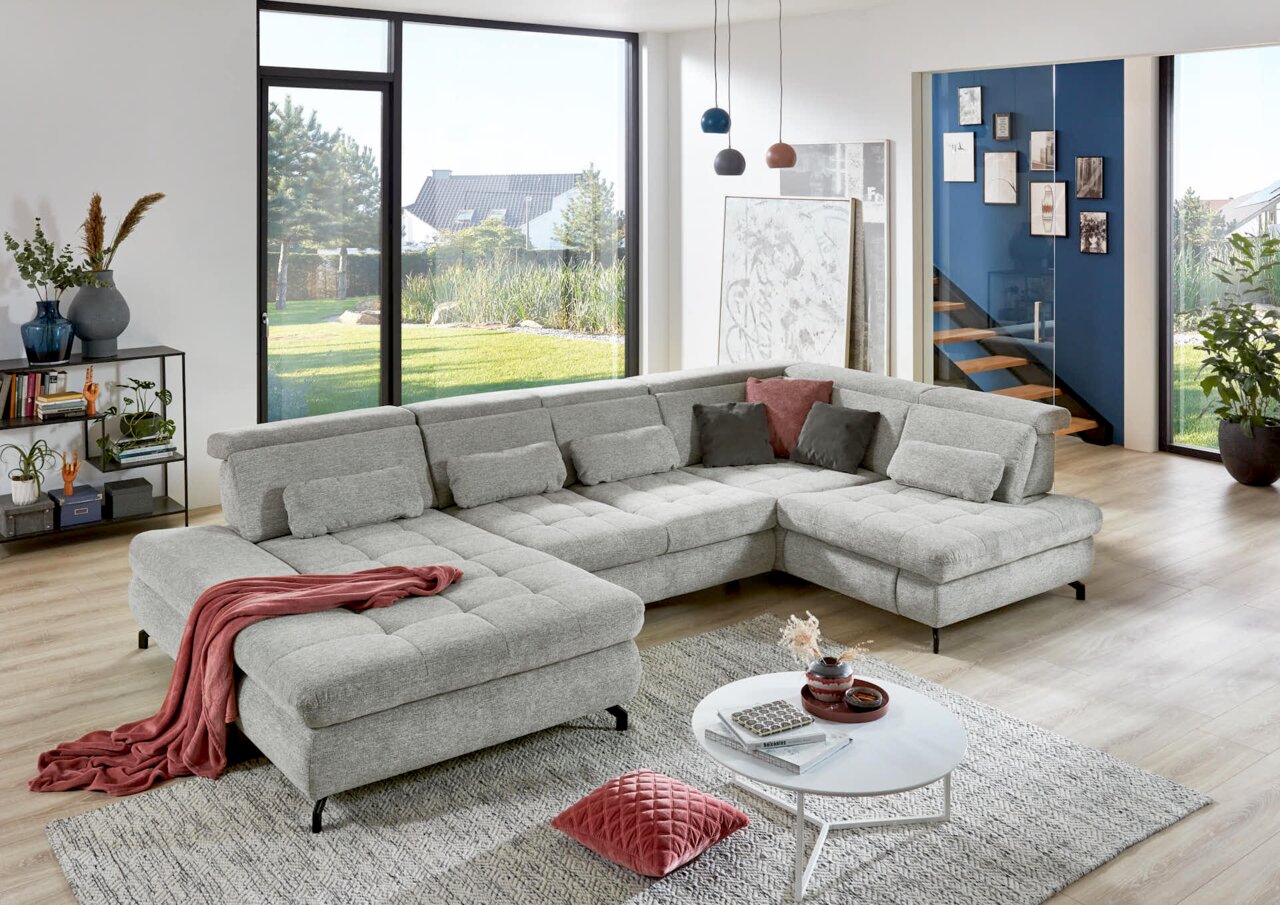 Graue Polsterrundecke in modernem Wohnzimmer mit großen Fenstern, dekoriert mit Kissen und Decke, seitliche Perspektive.