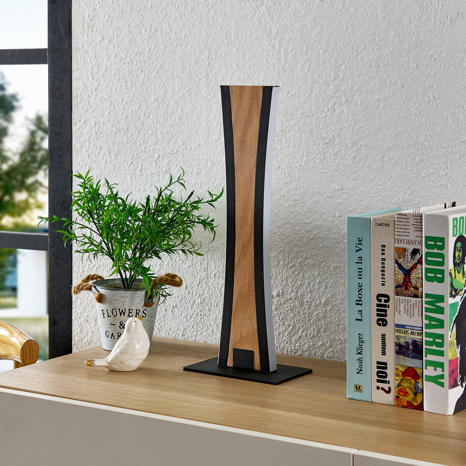 Moderne Tischlampe mit schlankem, geometrischem Design in Holzoptik und schwarzem Rahmen auf Sideboard neben Büchern und Pflanze, Frontansicht