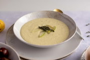 Cremige, hellgelbe Suppe in weißer Schale auf Teller, garniert mit Birnenscheiben und Thymianzweig; Aufnahme aus leicht erhöhter, schräger Vorderansicht.