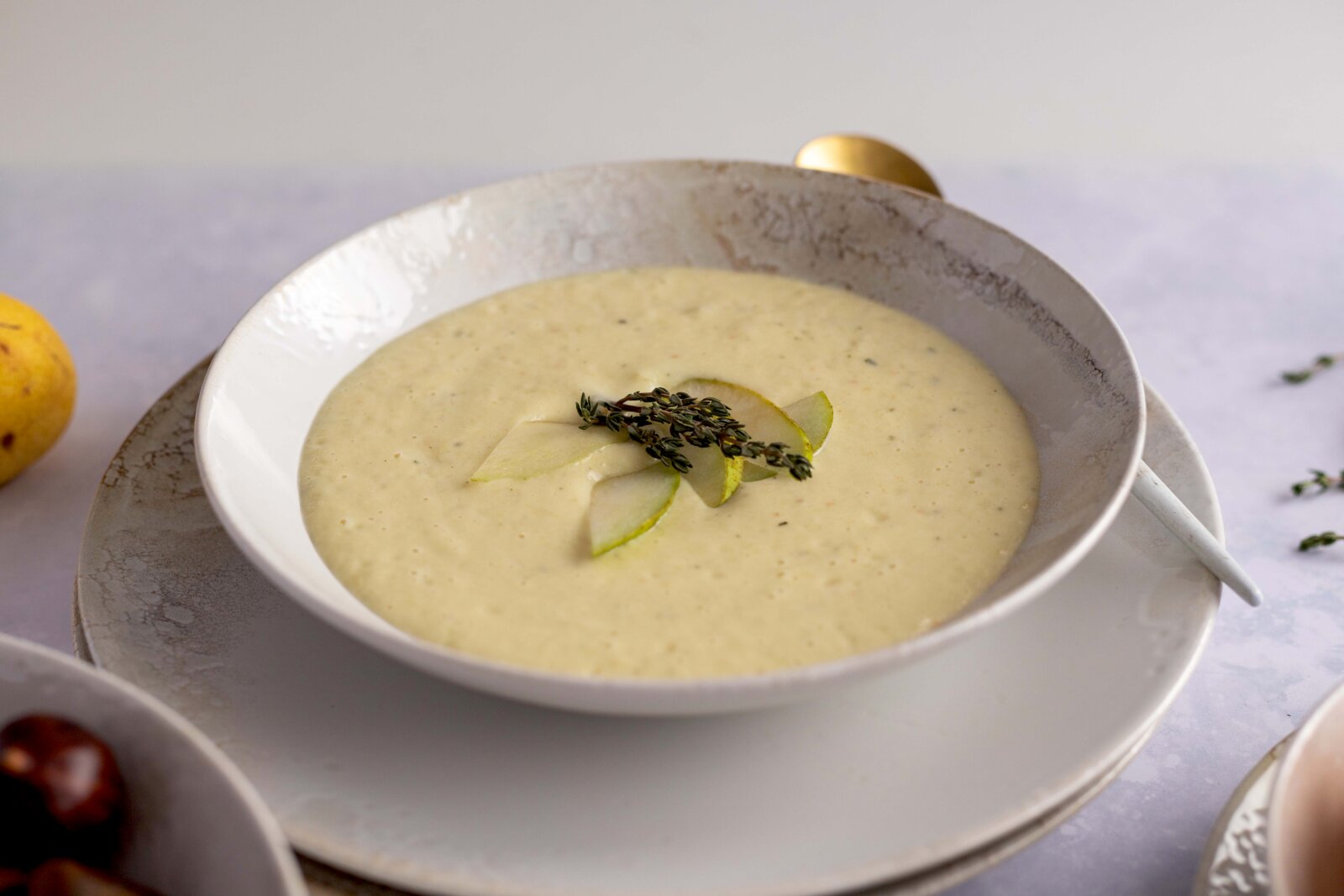 Cremige, hellgelbe Suppe in weißer Schale auf Teller, garniert mit Birnenscheiben und Thymianzweig; Aufnahme aus leicht erhöhter, schräger Vorderansicht.