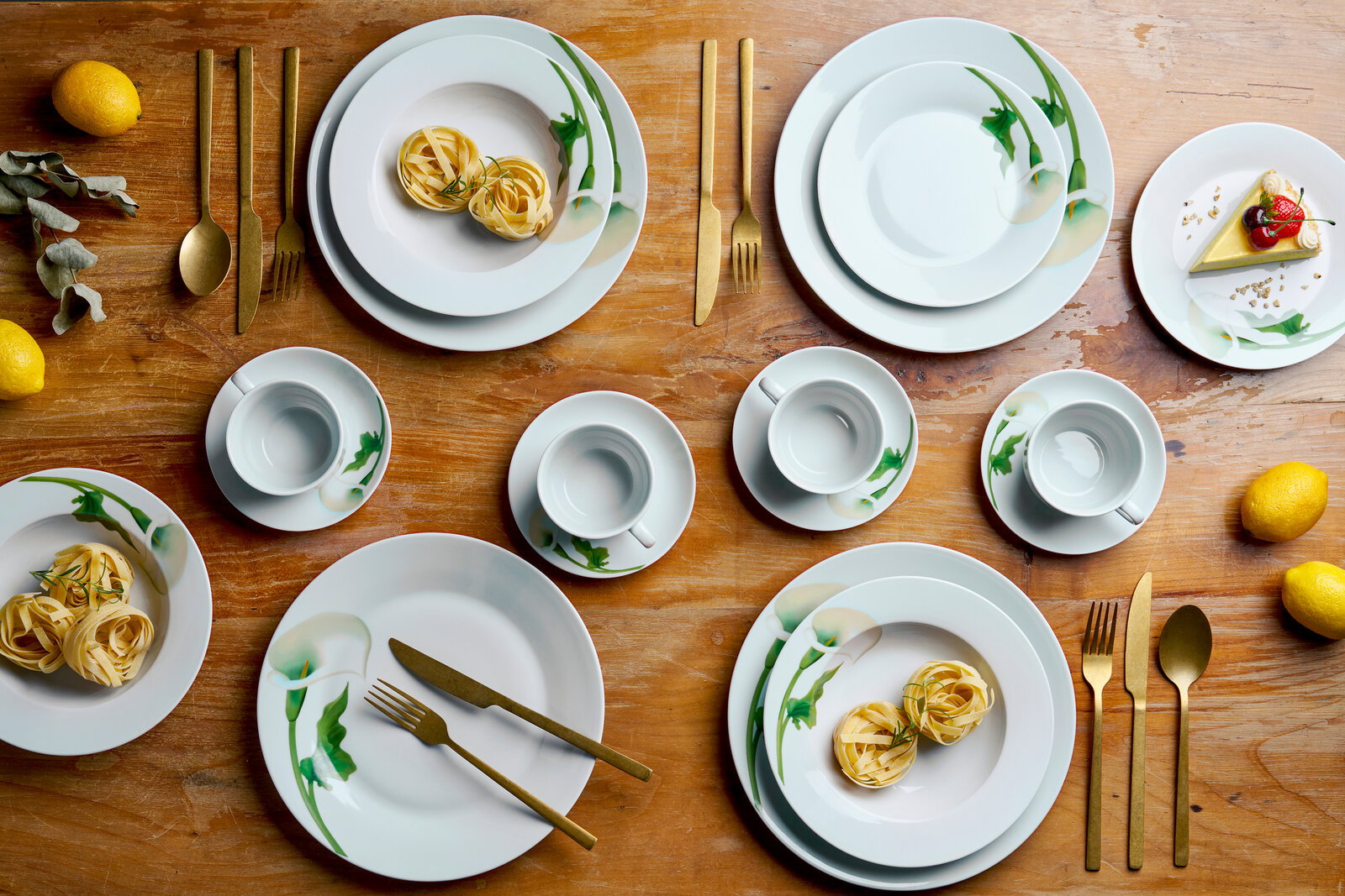 Draufsicht auf ein elegantes weißes Porzellan-Geschirrset mit grünem Blumenmuster (Teller und Tassen) auf Holztisch, arrangiert mit goldfarbenem Besteck, Zitronen und kleinen Speisen.