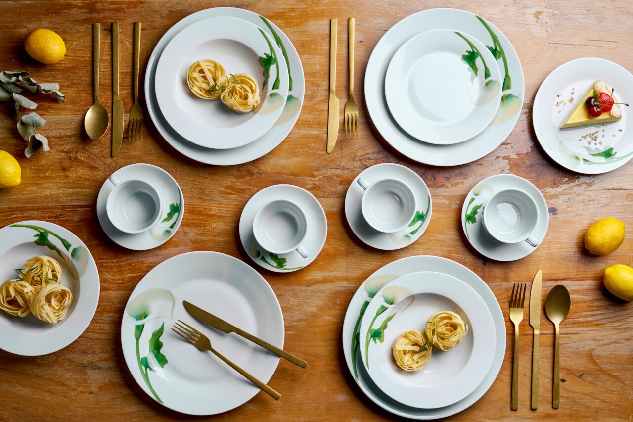 Draufsicht auf ein elegantes weißes Porzellan-Geschirrset mit grünem Blumenmuster (Teller und Tassen) auf Holztisch, arrangiert mit goldfarbenem Besteck, Zitronen und kleinen Speisen.
