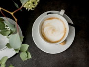 Draufsicht: Cappuccino mit Milchschaum in weißer Tasse auf Untertasse, mit kleinem Keks und Löffel auf dunkler Tischoberfläche, daneben grüne Blätter.