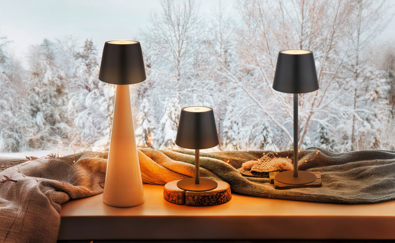 Drei Akku-Tischleuchten VANNIE 3 in verschiedenen Größen auf einer Fensterbank mit winterlicher Landschaft im Hintergrund, aus seitlicher Perspektive fotografiert.