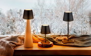 Drei Akku-Tischleuchten VANNIE 3 in verschiedenen Größen auf einer Fensterbank mit winterlicher Landschaft im Hintergrund, aus seitlicher Perspektive fotografiert.