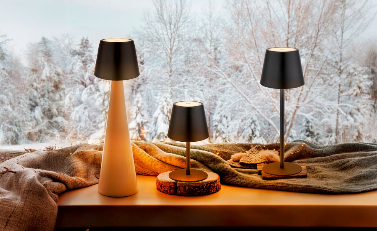 Drei Akku-Tischleuchten VANNIE 3 in verschiedenen Größen auf einer Fensterbank mit winterlicher Landschaft im Hintergrund, aus seitlicher Perspektive fotografiert.