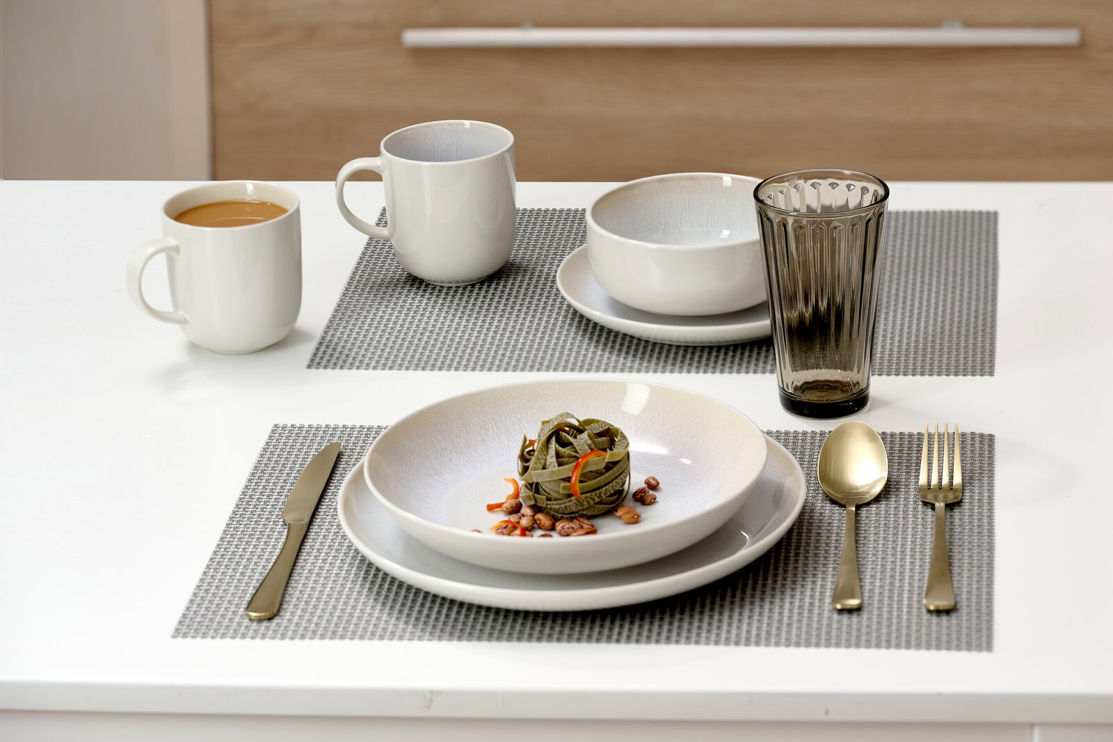 Tischgedeck mit zwei weißen Bechern, einer Schale, einem Glas und Besteck auf grauen Platzdeckchen, seitliche Perspektive.